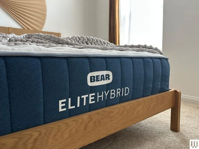Closeup of the front end of a Bear Elite Hybrid Mattress with blue trim and white top on a minimalist wooden bed frame