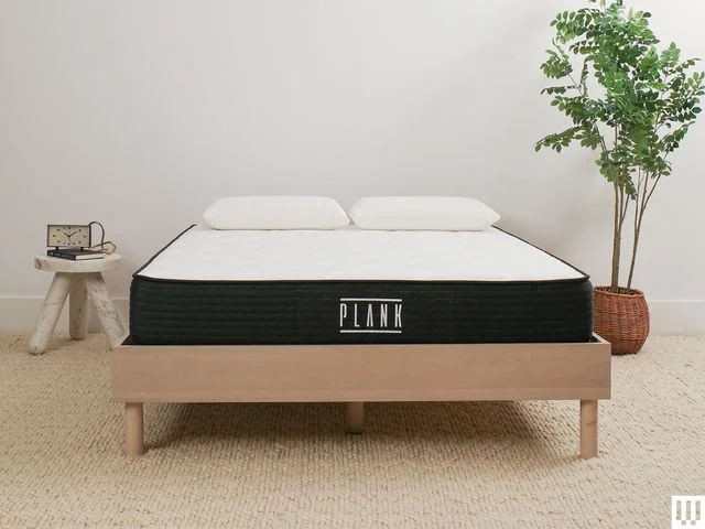 Plank Firm Mattress a white mattress with blue trim sitting on a minimalist wooden frame with a nightstand and potted...